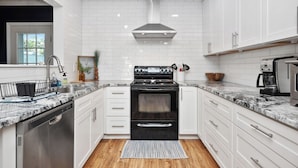A modern kitchen with white cabinetry, a black stove, and a stainless steel dishwasher. - Beach Please: Modern Coastal Bungalow Near Waterway and Marshwalk (Oak Island)