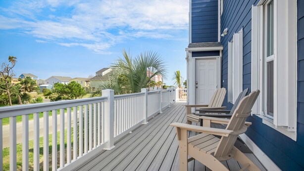 A wooden deck with two chairs and a table, overlooking a residential area with houses and greenery. - A Pier to Sea: Elegant Home with Private Pool, Hot Tub, Expansive Deck (Oak Island)