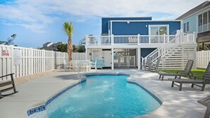 A swimming pool with a white deck, a two-story building with a blue exterior, and a white fence. - A Pier to Sea: Elegant Home with Private Pool, Hot Tub, Expansive Deck (Oak Island)