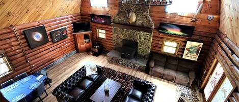 Interior d'una cabana de fusta amb una llar de pedra, un sofà de cuir negre, una taula de menjador amb caires i una televisió penjada.