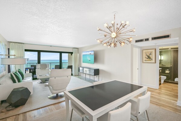A modern living room with a dining area, a large window with a view, a chandelier, and a television on the wall. - Huntington By The Sea 206. Gulf Front, Community Pool, Free Beach Service. (Miramar Beach)