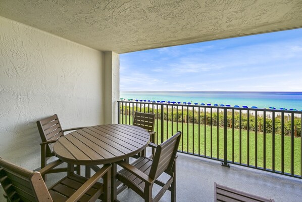 A balcony with a wooden table and chairs, a view of the ocean, and a green lawn.