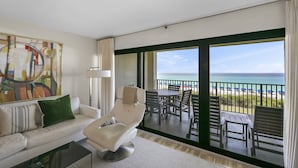 Un salon moderne avec un canapé, un fauteuil et un grand tableau. Il comprend un balcon offrant une vue sur la plage et l’océan.