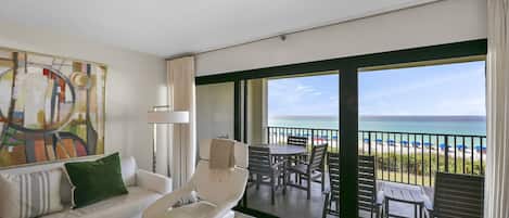 A modern living room with a sofa, armchair, and a large painting. It features a balcony with a view of the beach and ocean.