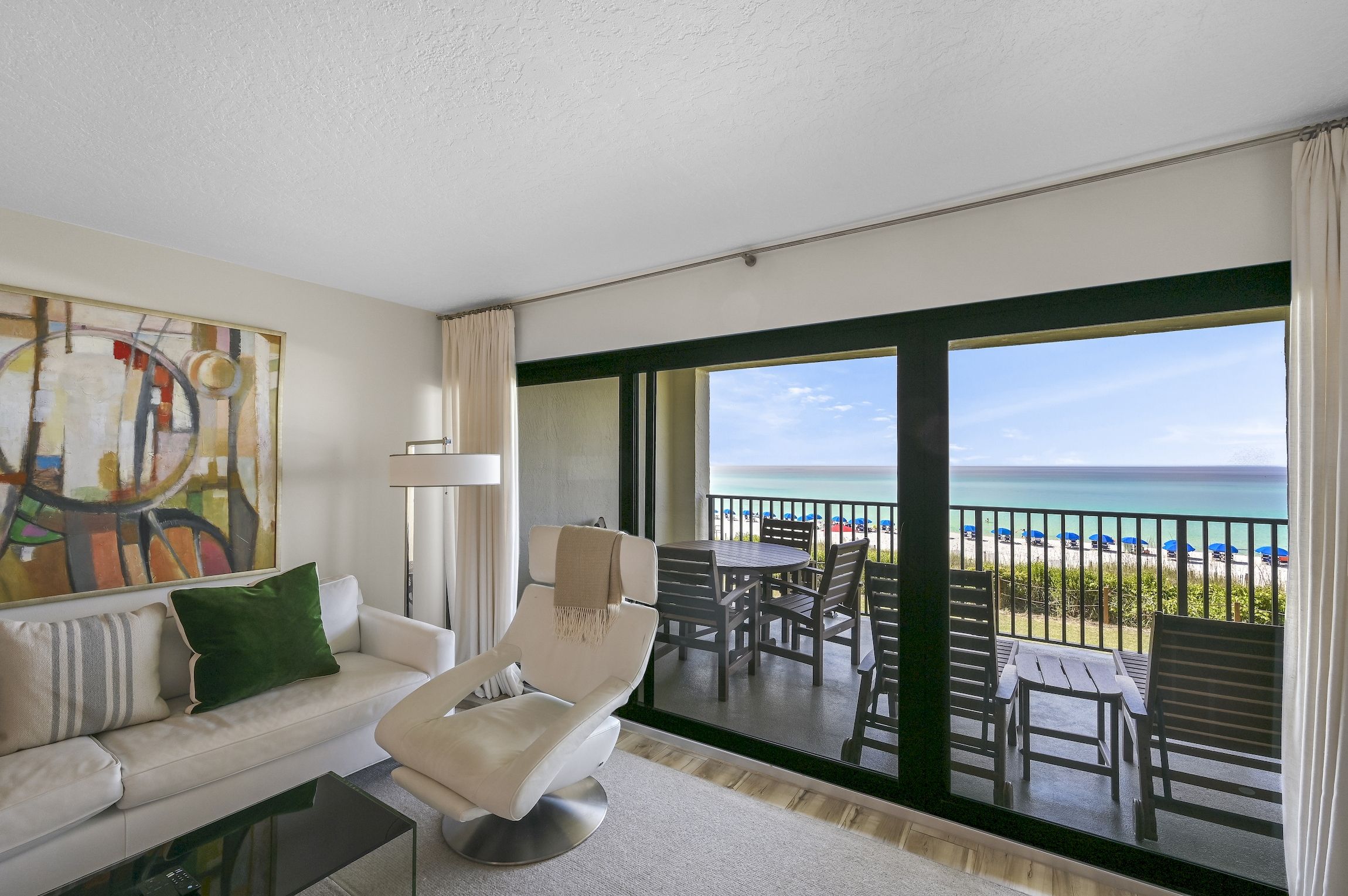 Un salon moderne avec un canapé, un fauteuil et un grand tableau. Il comprend un balcon offrant une vue sur la plage et l’océan.