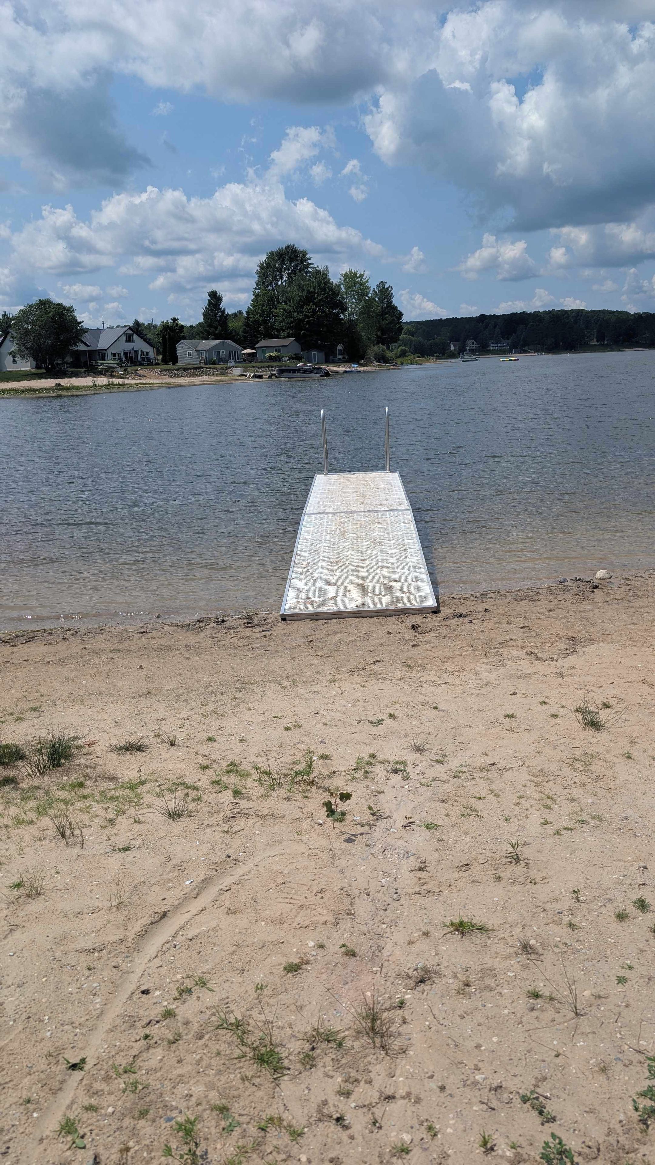 Une plage de sable avec une jetée métallique qui s’avance dans un lac paisible, entourée d’arbres et de maisons en arrière-plan.