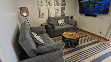 Un salon avec deux canapés gris, une table basse, un téléviseur fixé au mur et une enseigne portant l’inscription « VIE AU BORD DU LAC ».