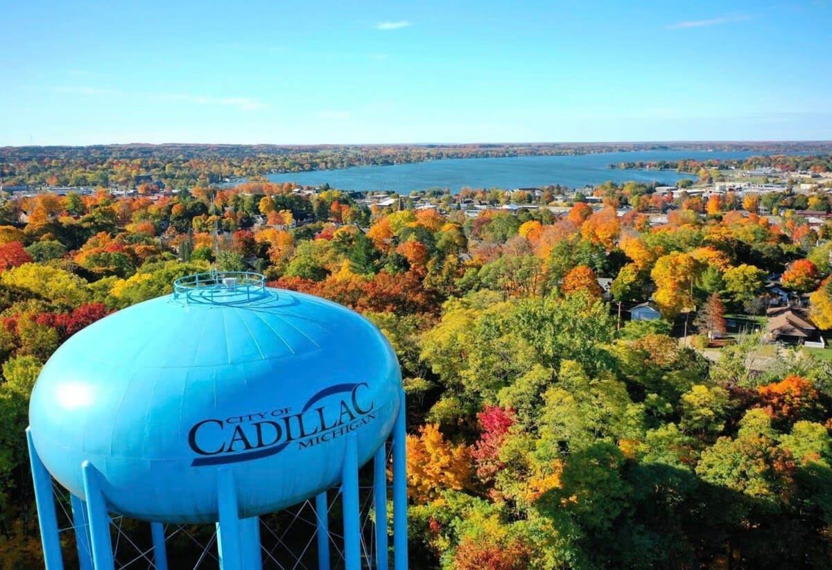 A water tower with 'City of Cadillac, Michigan' text, surrounded by autumn foliage and a lake view.