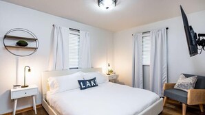 Ein modernes Schlafzimmer mit einem großen Bett, einem Nachttisch mit Lampe, einem an der Wand montierten Fernseher und einem Sessel.