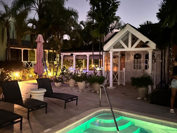 A pool area with lounge chairs, a covered seating area, and a small building with a white facade.