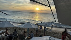 En strandscen med människor som sitter under parasoller, ett bord med drycker och en solnedgång över havet.