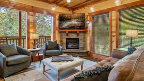 A cozy cabin interior with a stone fireplace, wooden walls, and large windows. - Pool! Hot Tub - Movie Room - Game Room - Dollywood (Sevierville)