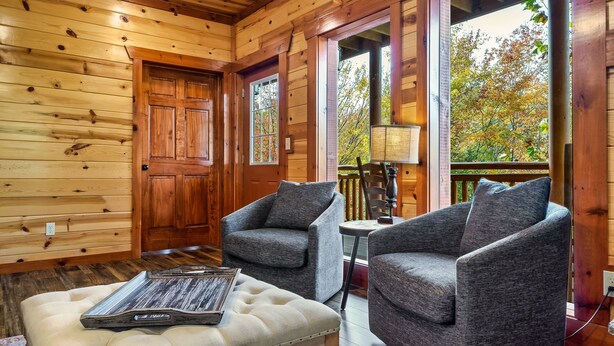 A cozy cabin interior with wooden walls, two grey armchairs, a white cushioned bench, and a wooden door. - Pool! Hot Tub - Movie Room - Game Room - Dollywood (Sevierville)