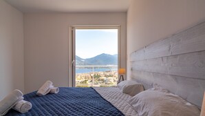 A hotel room with a large bed, a view of a city and mountains through a window, and a wooden headboard. - Villa A Cucina (Propriano)
