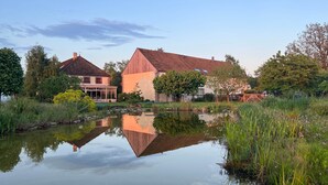 A serene lakeside setting with a wooden house and a smaller building, surrounded by lush greenery and a calm pond reflecting the structures. - LA MELINOTHE (Paisy-Cosdon)