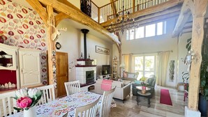 A spacious living area with a high ceiling, wooden beams, a fireplace, and a dining area with a table and chairs. - LA MELINOTHE (Paisy-Cosdon)
