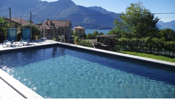 Ein Swimmingpool mit klarem, blauem Wasser, umgeben von einer Terrasse mit Liegestühlen und einem kleinen Gebäude. Im Hintergrund sind Berge und ein Gewässer zu sehen.