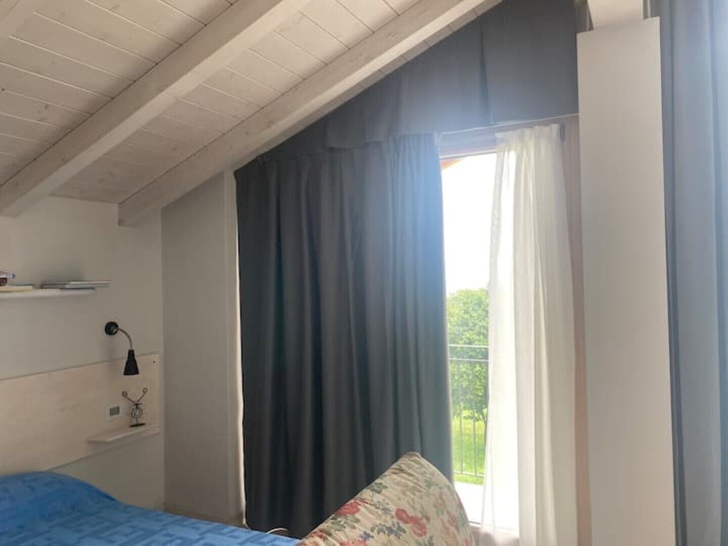 Een slaapkamer met een bed, een raam met gordijnen, een plank met een lamp en uitzicht op groen.