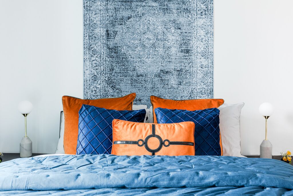 A bed with a blue quilted cover, two orange pillows, and a textured wall tapestry.