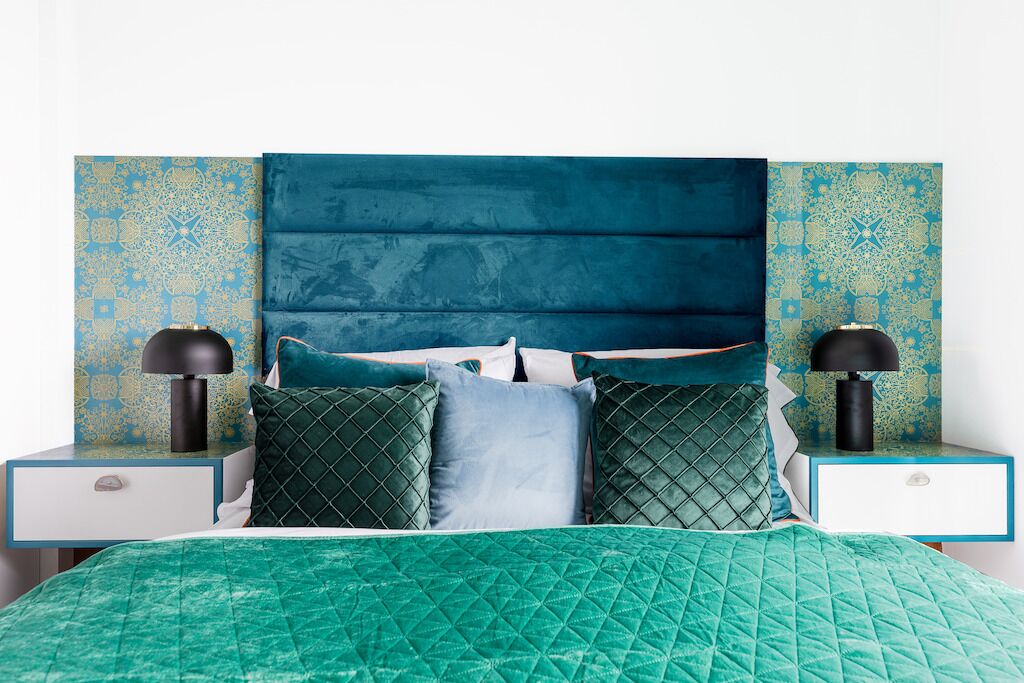 Un lit recouvert d’une courtepointe turquoise à motifs matelassés, avec une tête de lit rembourrée en tissu bleu et des coussins assortis. On y trouve deux tables de chevet avec des lampes et un panneau décoratif mural.