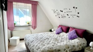 Une chambre à coucher avec un lit, une fenêtre ornée de rideaux roses, un radiateur et un mur sur lequel figure une inscription décorative.