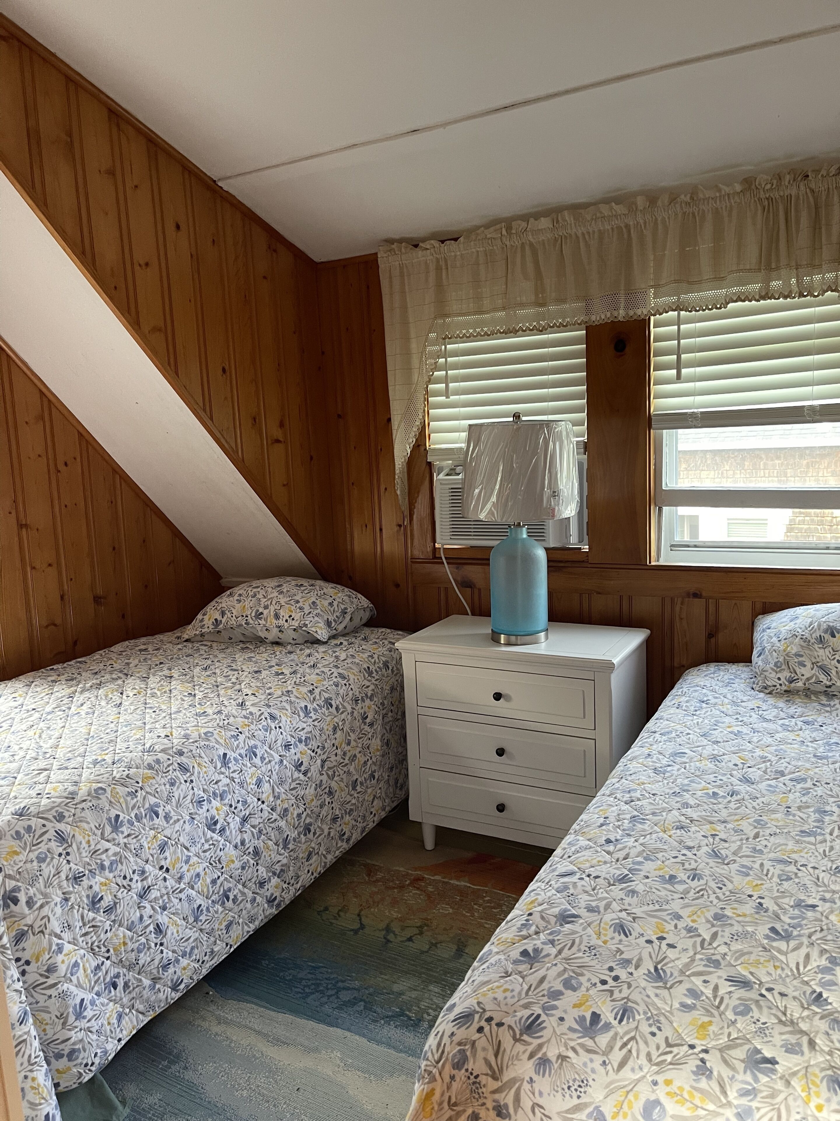 Une chambre avec deux lits, une table de chevet, une lampe et une fenêtre avec des stores.