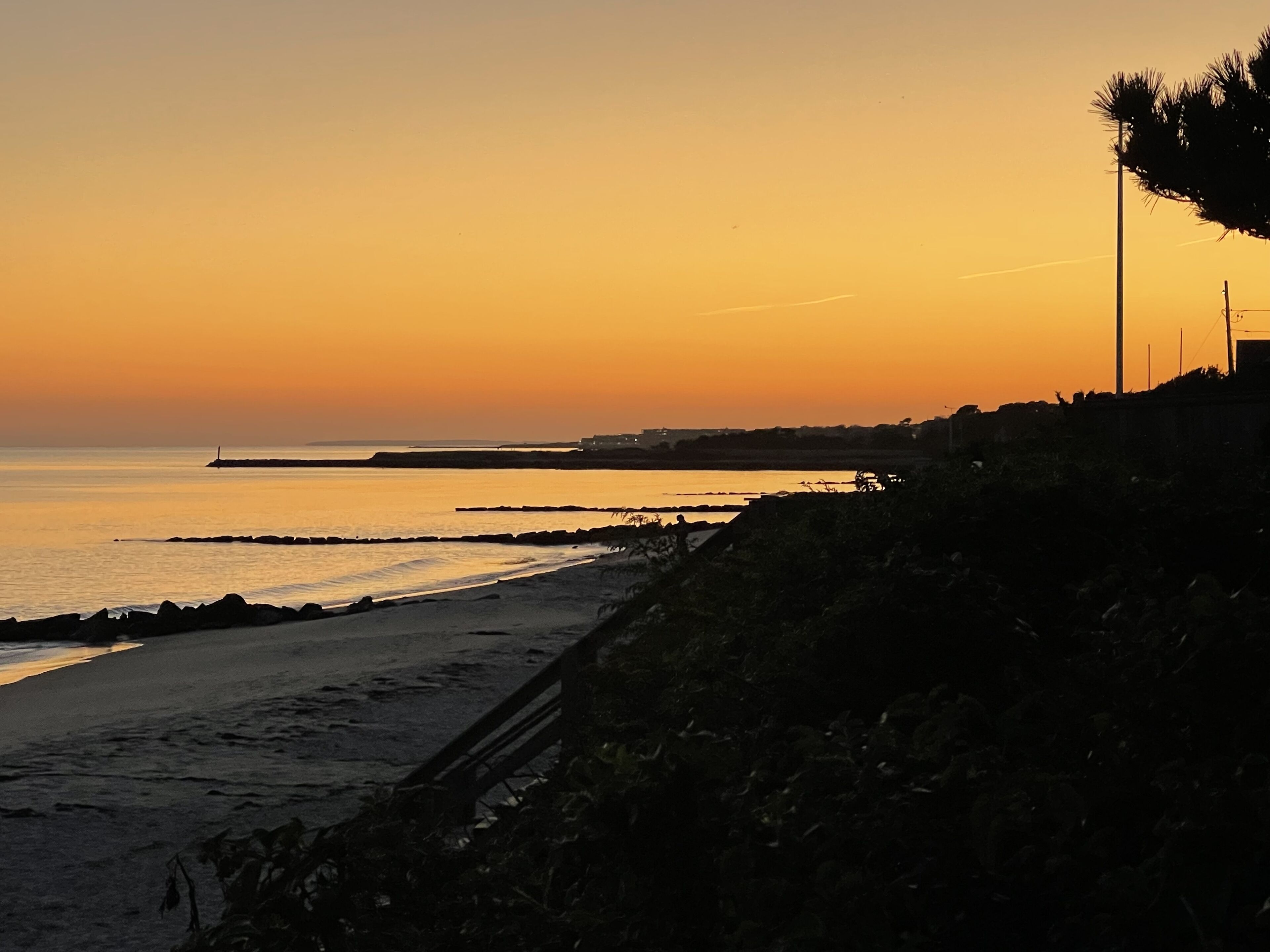 A coastal sunset with a beach, rocky shoreline, and a lighthouse in the distance.