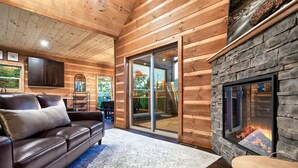 Ein gemütliches Blockhaus-Interieur mit einem Ledersofa, einem Kamin aus Naturstein und einer großen Schiebeglastür, die zu einem Außenbereich führt.