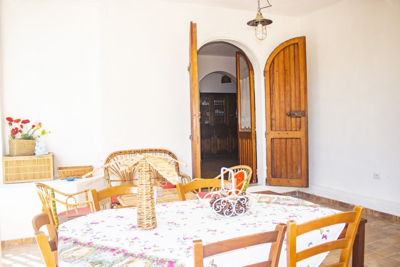 Un coin repas avec une table ronde, des chaises en osier et une nappe à motifs floraux. On y trouve une porte en bois et une arche ouverte qui mène à une autre pièce.