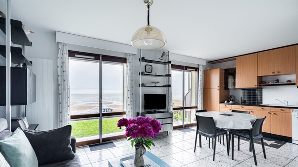 Ein modernes Wohnzimmer mit einer Couch, einem Couchtisch mit Blumenvase und einem Essbereich mit Tisch und Stühlen. Es gibt eine Küchenzeile mit Schränken und einem Kühlschrank. Große Fenster bieten Blick auf den Ozean.