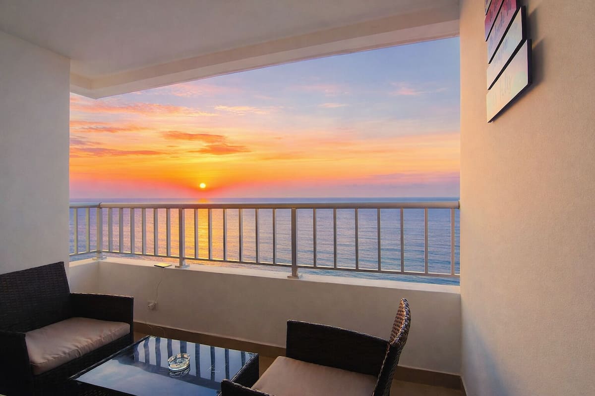 Un balcon offrant une vue sur le coucher de soleil sur l’océan, aménagé avec deux chaises et une petite table.