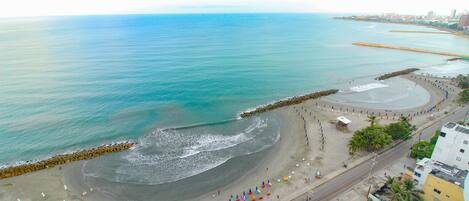 An aerial view of a beach with a road running parallel to the shore, a breakwater, and buildings in the background.