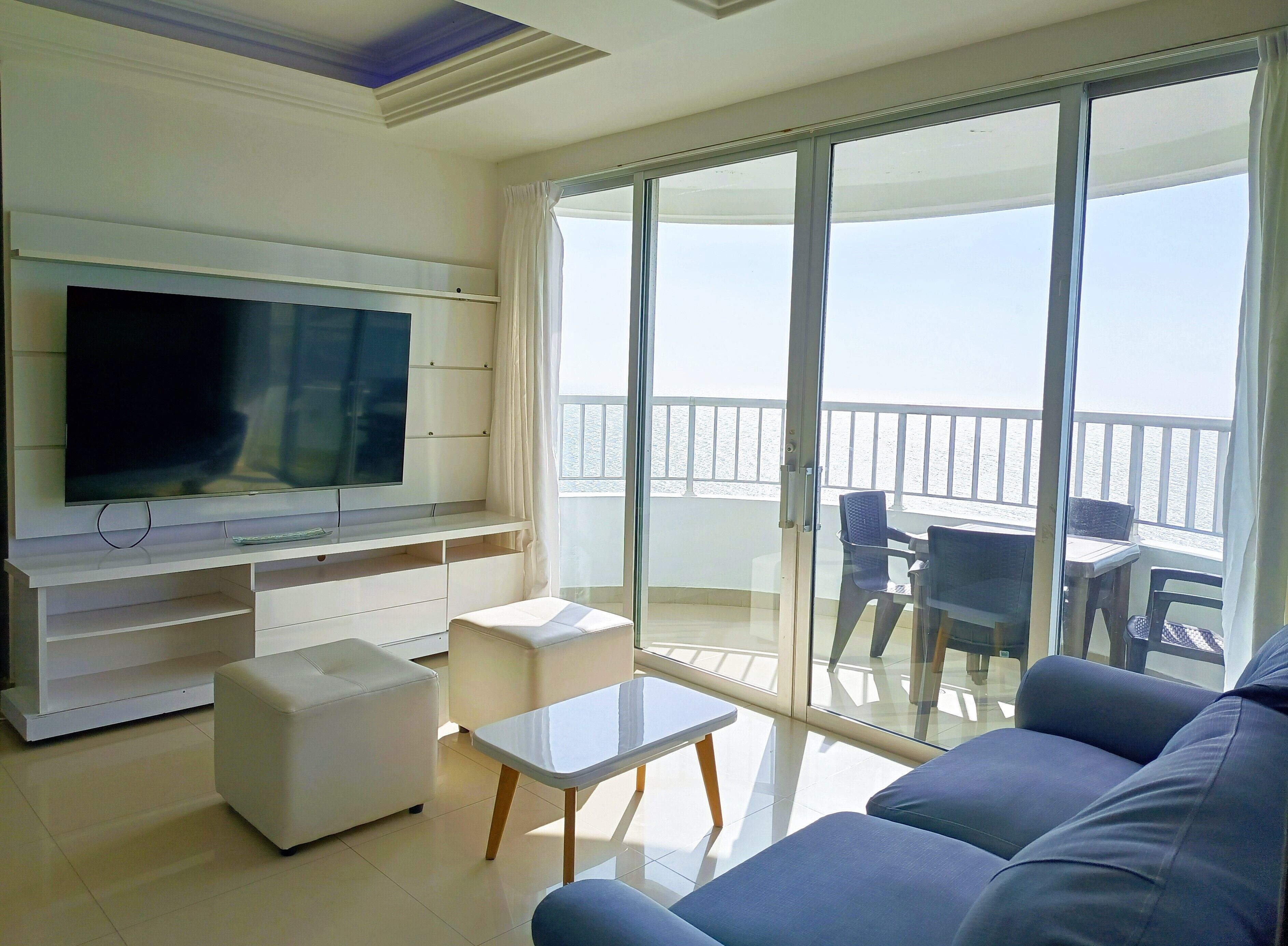 A modern living room with a flat-screen TV, a sofa, a coffee table, and a balcony with a view of the sea.