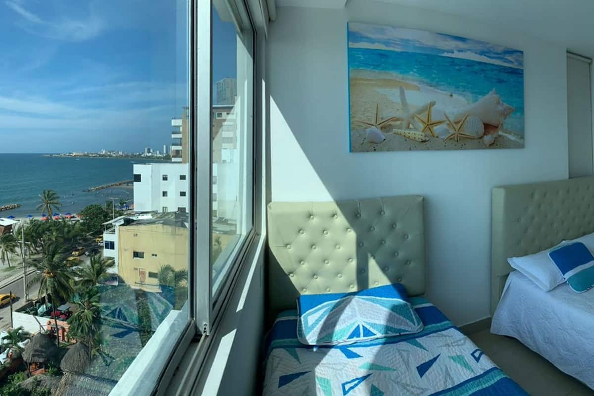 Une chambre d’hôtel avec un lit, une fenêtre donnant sur la mer et un tableau représentant une scène de plage.