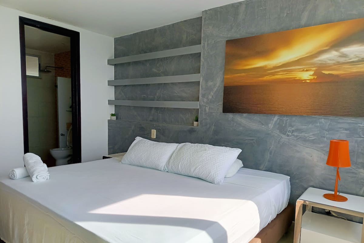 Une chambre moderne avec un grand lit, une table de chevet, un tableau représentant un coucher de soleil et une salle de bain visible à travers une porte ouverte.