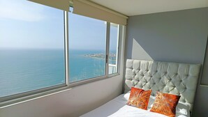 A bedroom with a large window, a bed with white bedding and red accents, and a tufted headboard. - Apartamentos | Frente al Mar + Primera Linea de Playa | Piscina TOP (Cartagena)