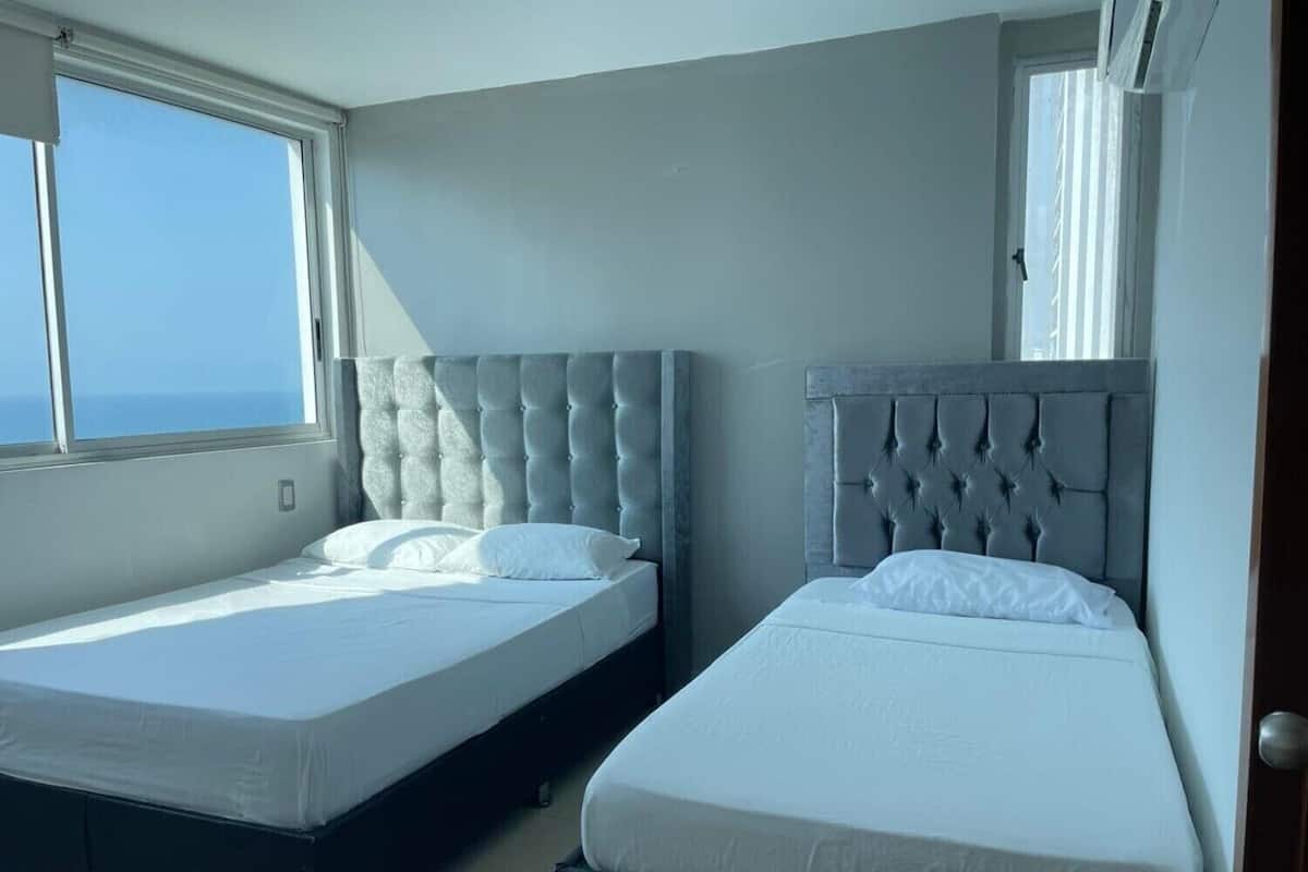 Une chambre à coucher avec deux lits, chacun doté d’une tête de lit capitonnée, de linge de lit blanc et offrant une vue sur l’océan par une fenêtre.