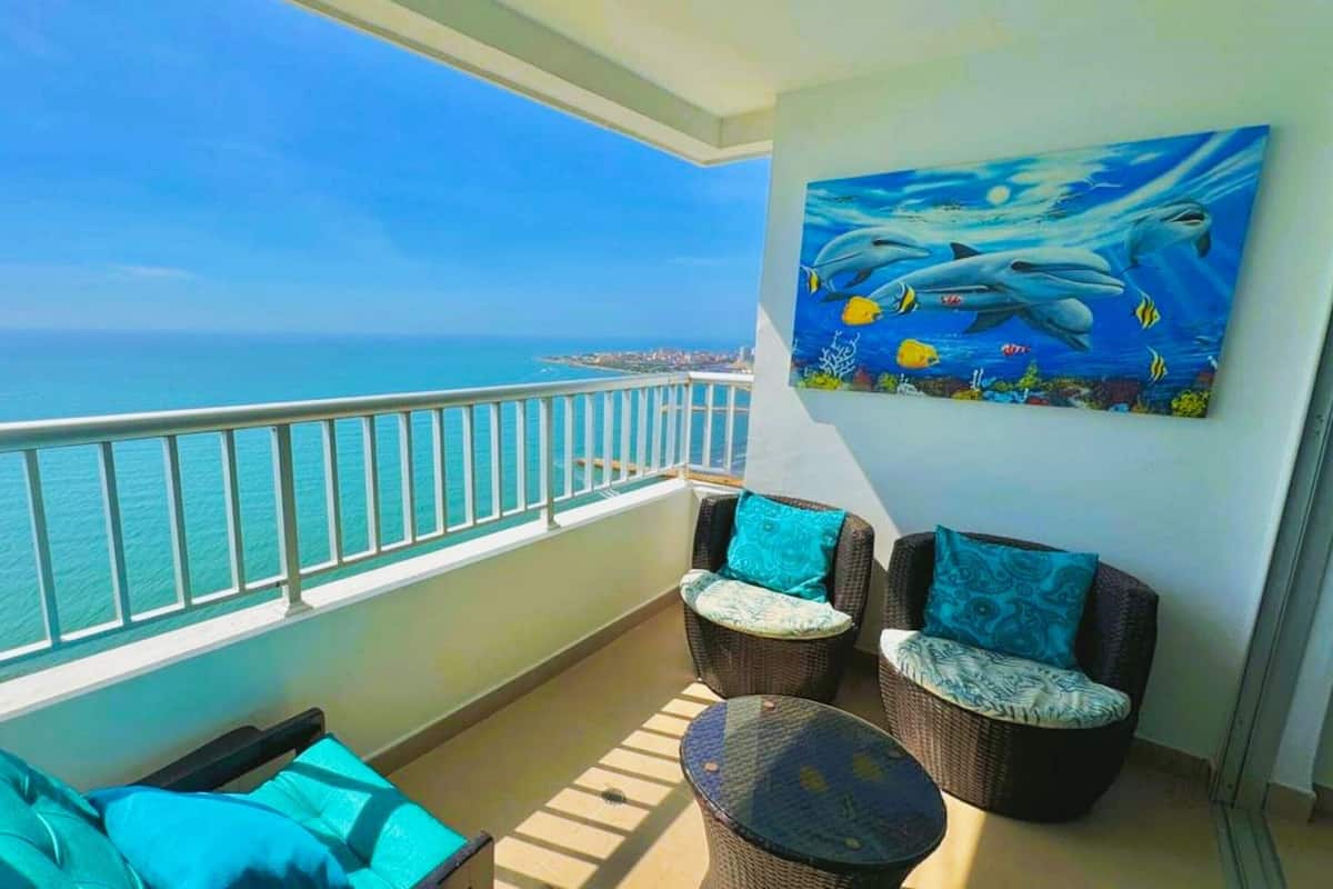 Un balcon avec vue sur l’océan, des meubles en osier et un tableau représentant des dauphins.