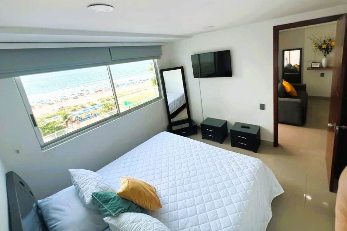 Une chambre à coucher avec un lit, une télévision, une fenêtre donnant sur la plage et une porte menant à une autre pièce.