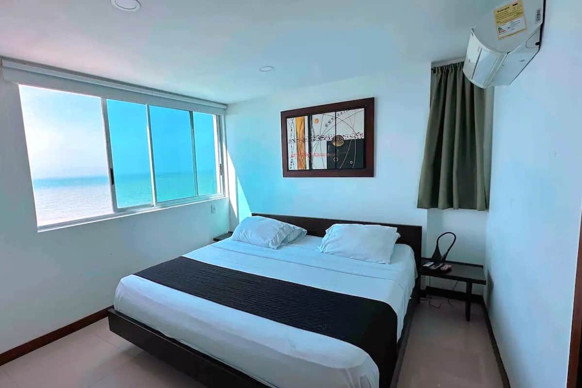 Une chambre d’hôtel avec un lit, une fenêtre donnant sur la mer, un tableau au mur et une table de chevet avec une lampe.