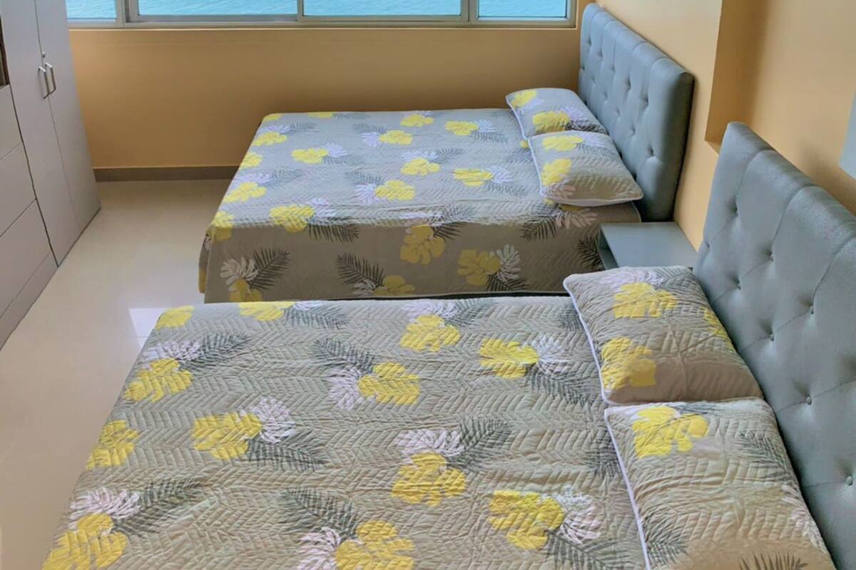 Une chambre dotée de deux lits, chacun recouvert d’une couvre-lit à motifs floraux, avec une tête de lit rembourrée de couleur grise et une grande fenêtre donnant sur la mer.