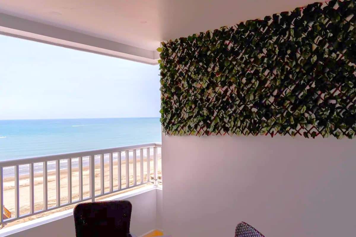 Une pièce dotée d’une grande fenêtre donnant sur une plage, d’une balustrade blanche et d’un mur recouvert de feuillage verdoyant.