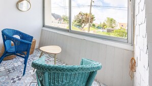 Een kamer met een groot raam, twee rieten stoelen, een kleine ronde tafel en een spiegel aan de muur.