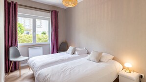 Una habitación de hotel con una cama grande, una silla, una mesita de noche con lámpara y una ventana con cortinas.