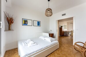 Une chambre à coucher avec un lit, une table de chevet, une lampe, un fauteuil en osier et un couloir menant à une autre pièce.