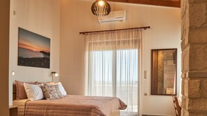 Une chambre à coucher comprenant un lit, une table de chevet, un climatiseur fixé au mur, une grande fenêtre avec des rideaux légers et un tableau représentant un coucher de soleil.