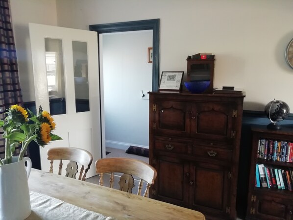 A dining area with a wooden table, two chairs, a vase with sunflowers, a wooden cabinet with a globe and books, and a door with a mirror. - Cosy, Spacious 2-bed Cottage in Watchet, Somerset (Watchet)