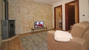 Uma sala de estar com parede de pedra, lareira, televisão, sofá e porta que dá para outro cômodo.