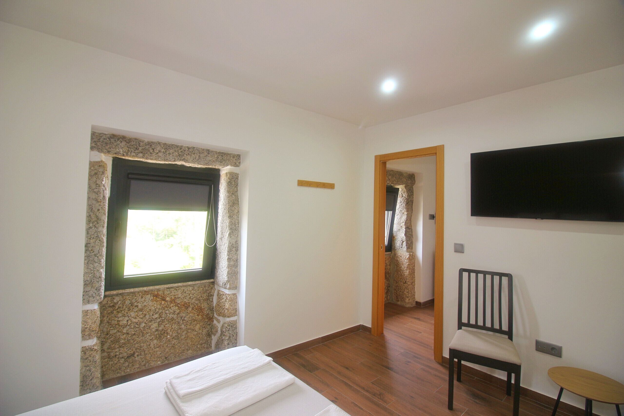 A bedroom with a stone wall, a bed, a chair, a small table, and a TV mounted on the wall.