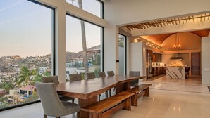 A modern dining area with a large wooden table and benches, large windows offering a city view, and a kitchen visible in the background. - Luxury Villa w/ Tennis, Pickleball, Pool, Hot Tub & Fitness Room, Views (Cabo San Lucas)
